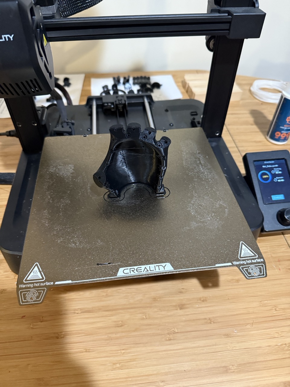 Completed wrist connector piece on print bed with printer screen showing print progress, other arm components laid out in background