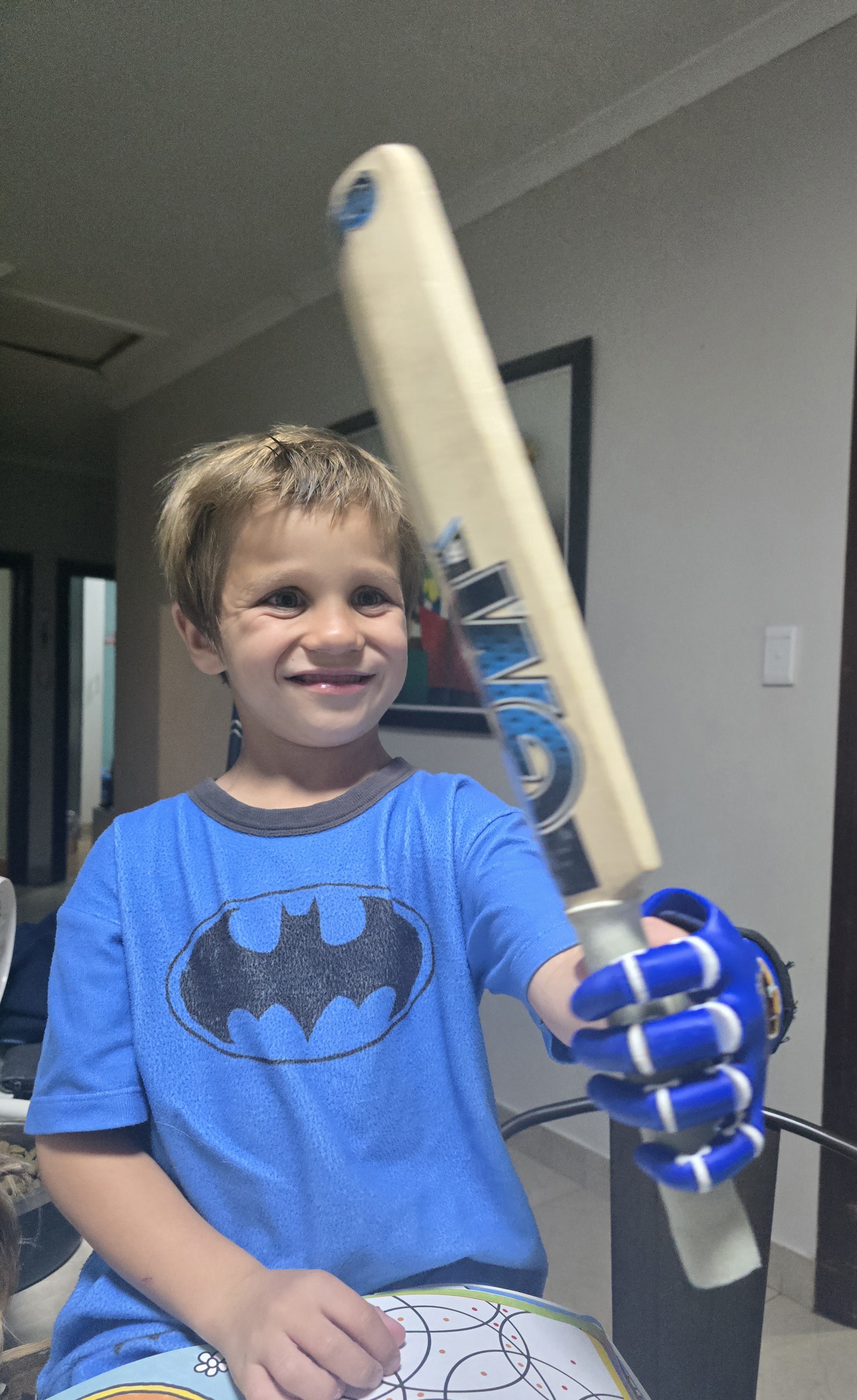 Noah smiling and holding a cricket bat with his dark blue prosthetic hand, blue Batman t-shirt, Strand South Africa