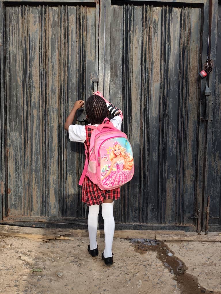 O.C. from behind in school uniform, reaching up with her prosthetic arm to open the school gate